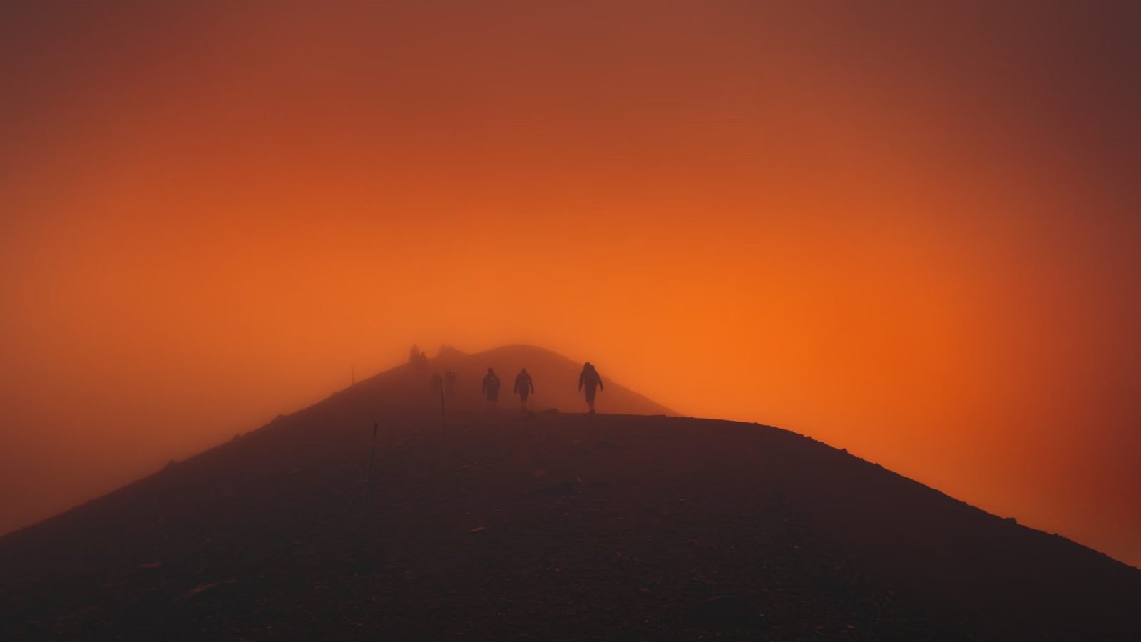 A line of hikers silhouetted against a vivid orange sunset, one leading the way along a mountain ridge
